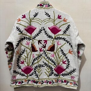 May include: A light-colored jacket with an embroidered floral design. The jacket features a cream-colored base with a detailed pattern of pink, orange, and green flowers and leaves. The embroidery is visible on the collar, sleeves, and hem.