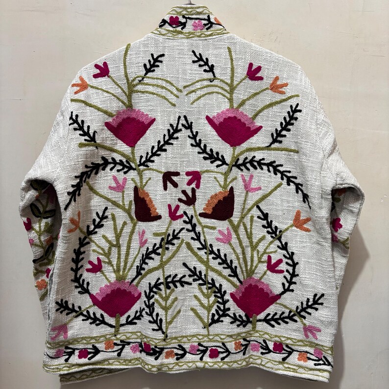 May include: A light-colored jacket with an embroidered floral design. The jacket features a cream-colored base with a detailed pattern of pink, orange, and green flowers and leaves. The embroidery is visible on the collar, sleeves, and hem.