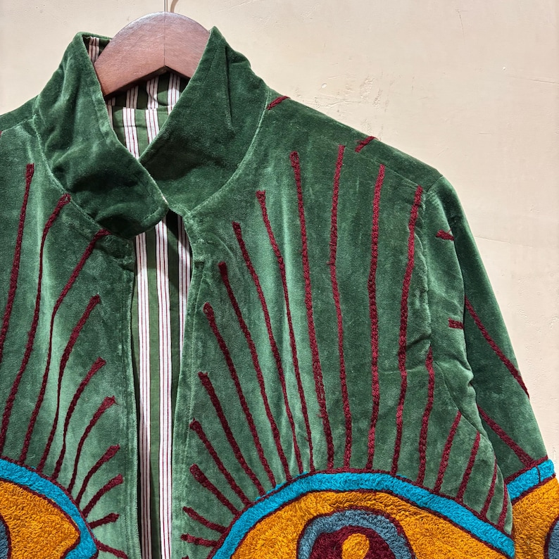 May include: Green velvet jacket with embroidered sunburst design in red, yellow, and blue. The jacket has a striped lining and a collared neckline. The jacket is hanging on a wooden hanger.