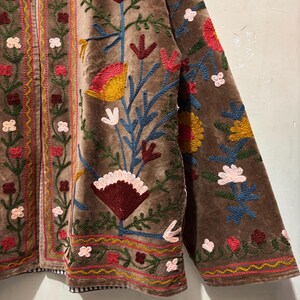 May include: Brown velvet jacket with colourful floral embroidery. The jacket features a detailed design with red, yellow, blue, and white flowers and leaves. The embroidery is on the front, sleeves, and edges of the jacket.