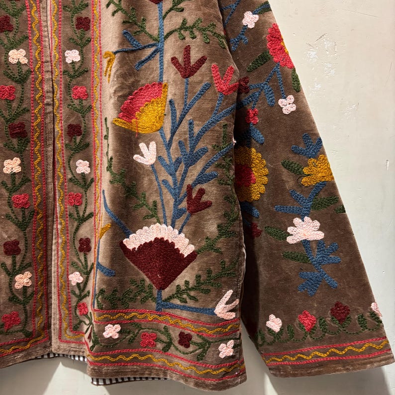 May include: Brown velvet jacket with colourful floral embroidery. The jacket features a detailed design with red, yellow, blue, and white flowers and leaves. The embroidery is on the front, sleeves, and edges of the jacket.
