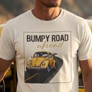 May include: A cream-colored t-shirt with the text "Bumpy Road Ahead" and a graphic of a yellow car on a road. The person is standing next to a yellow vintage car with the door open. The background features a scenic view of mountains and the ocean.
