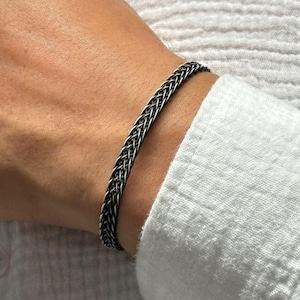 May include: A silver-toned bracelet featuring a detailed, intricate woven chain design. The metal has a darkened, oxidized finish within the crevices of the weave, creating a high-contrast appearance that highlights the complex braided texture.