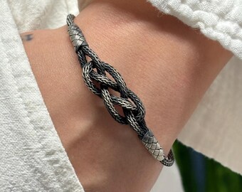 May include: A silver bracelet featuring a central knot design with a dark, oxidized finish. The bracelet consists of a woven, rope-like chain that transitions into smooth, textured silver end caps on both sides of the central knot element.