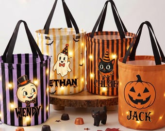 Op de afbeelding: Vier cilindervormige Halloween-snoepemmers met zwarte hengsels en interne ledverlichting. De emmers hebben verticale strepen in paars, geel en oranje, versierd met illustraties van een geest, kat, pompoen en personage, gepersonaliseerd met de namen Wendy, Ethan, Chloe en Jack.