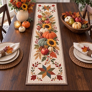 May include: A rectangular quilted table runner featuring a central autumn-themed print of pumpkins, sunflowers, wheat, and fall leaves, framed by geometric star patterns at both ends. The runner is displayed on a dark wood table with place settings.