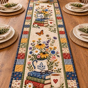 May include: A rectangular quilted table runner featuring a cream-colored center panel with printed illustrations of stacked books, floral pitchers, butterflies, and mugs with heart motifs, bordered by a patchwork of colorful floral and checkered fabric squares.