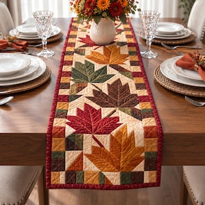 May include: A rectangular quilted table runner featuring a patchwork design of autumn maple leaves in shades of red, orange, yellow, brown, and dark green. The runner has a border of small squares and is centered on a wooden dining table.
