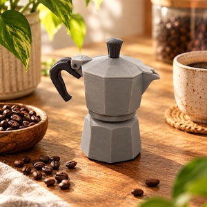 Könnte beinhalten: Eine graue Mokkakanne mit facettiertem, achteckigem Körper, einem dunkelgrauen Griff und einem passenden dunkelgrauen Knopf auf dem Deckel. Die Kaffeebereiter weist eine sichtbare Schichtstruktur auf, die für 3D-Drucke typisch ist, und steht aufrecht.