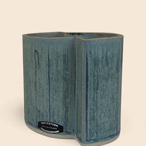 Op de afbeelding: Een turquoise keramische vaas met een uniek, gebogen ontwerp. De vaas heeft een getextureerd oppervlak en een label met de tekst "Pottestuen Handmade in Denmark Frederikshavn."