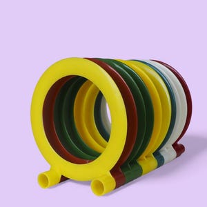 Può includere: Un giocattolo ad anelli multicolore con anelli gialli, rossi, verdi e bianchi. Il giocattolo è composto da diversi anelli circolari collegati alla base. Lo sfondo è di colore lavanda uniforme. Per giocare o decorare.