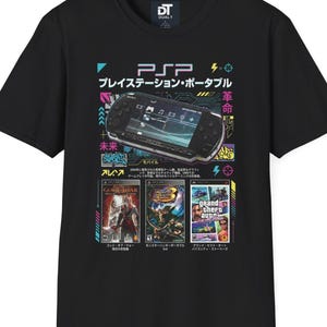 Może przedstawiać: Czarny t-shirt z grafiką konsoli PSP, japońskim tekstem i okładkami gier. Projekt zawiera tekst "PSP プレイステーション・ポータブル" i tytuły gier. Koszulka ma okrągły dekolt i krótkie rękawy.