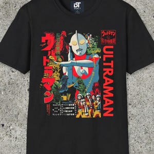Puede incluir: Camiseta negra con un gráfico colorido de Ultraman, un superhéroe japonés, y texto japonés. El diseño incluye la palabra "ULTRAMAN" en rojo, junto con imágenes de monstruos y un equipo con uniformes amarillos.