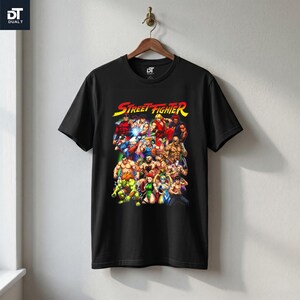 May include: A black short-sleeve t-shirt featuring a colorful graphic print of various Street Fighter video game characters. The text Street Fighter is printed in yellow and red at the top of the graphic. The shirt has a crew neckline.