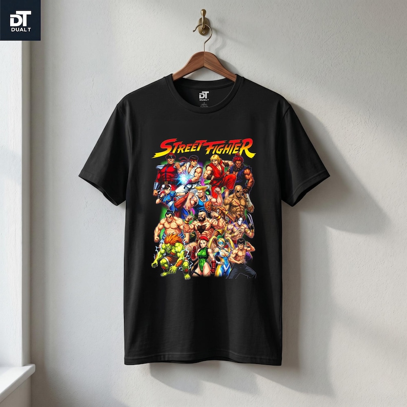 May include: A black short-sleeve t-shirt featuring a colorful graphic print of various Street Fighter video game characters. The text Street Fighter is printed in yellow and red at the top of the graphic. The shirt has a crew neckline.