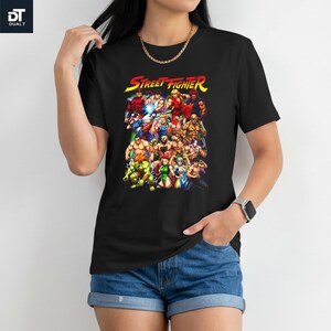 May include: A black short-sleeve t-shirt featuring a colorful graphic print of various Street Fighter video game characters. The text "STREET FIGHTER" is printed in yellow and red at the top of the graphic, which displays a group of fighters.