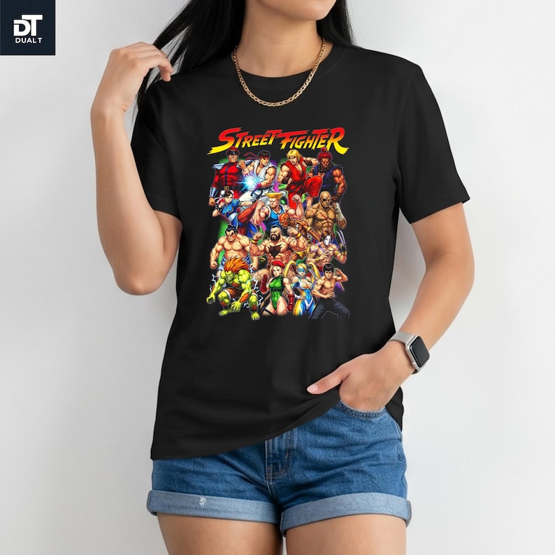 May include: A black short-sleeve t-shirt featuring a colorful graphic print of various Street Fighter video game characters. The text "STREET FIGHTER" is printed in yellow and red at the top of the graphic, which displays a group of fighters.