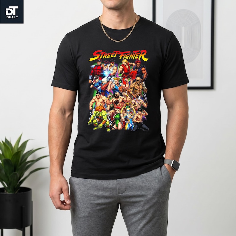 May include: A black short-sleeved t-shirt featuring a colorful graphic print of various Street Fighter video game characters arranged in a group. The text Street Fighter is printed in a stylized yellow and red font above the character illustration.