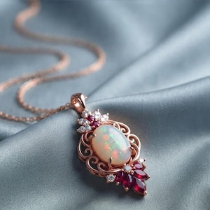 May include: A rose gold pendant necklace featuring a central oval cabochon opal with iridescent flashes. The setting is adorned with deep red marquise-cut gemstones and small clear accent stones arranged in a decorative filigree metal frame.