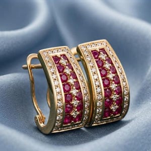 May include: A pair of gold hoop earrings featuring a curved rectangular design. Each earring is set with a central row of round, deep red rubies flanked by two rows of small, clear white diamonds, secured with a hinged post closure mechanism.