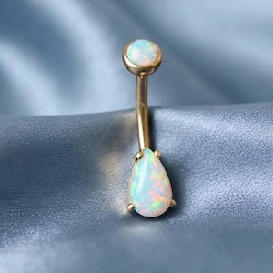 Peut inclure: Un piercing de nombril en or courbé avec un cabochon d'opale rond sur le dessus et un cabochon d'opale en forme de poire plus grand en bas. Les opales présentent des éclats irisés bleus, verts et roses sur une base blanche laiteuse.