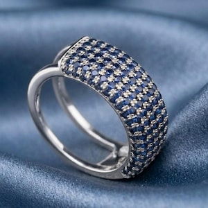 May include: A wide silver-toned ring featuring a curved band encrusted with multiple rows of small, round, deep blue stones. The band splits into two separate, thinner silver-toned shanks that meet at the base of the stone-covered section.