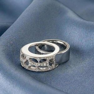 May include: A silver-toned ring featuring a wide band with an intricate, openwork lattice design embedded with small, clear stones. The ring is positioned partially overlapping a second, plain, smooth silver-toned band, creating a layered appearance.