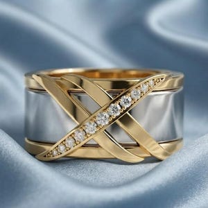 Könnte beinhalten: Ein breiter Ring mit einer polierten silberfarbenen Basis, die mit zwei sich kreuzenden goldfarbenen Bändern überlagert ist. Eines der goldenen Bänder ist mit einer Reihe kleiner, klarer, rund geschliffener Steine besetzt, die diagonal verlaufen.