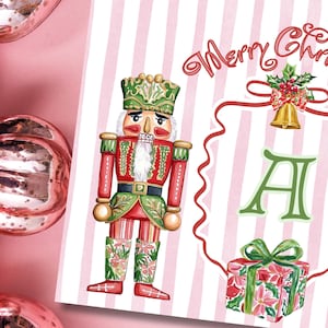 以下が含まれることがあります： ピンクと白のストライプの背景を持つ水彩画のクリスマスカード。カードには、赤と緑のくるみ割り人形の兵士、ギフトボックス、そして「メリークリスマス」という言葉が描かれています。ピンクのクリスマスオーナメントが背景にあります。