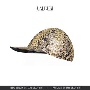 May include: A baseball cap crafted from genuine snake leather. The cap has a black peak and a beige and brown snakeskin pattern. The CALDERI logo is visible in the upper left corner. The bottom of the image reads "100% GENUINE SNAKE LEATHER".
