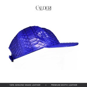 May include: A striking blue baseball cap crafted from genuine snake leather. The cap has a curved peak and a textured, scale-like design. The brand name "CALDERI" is displayed at the top. The bottom of the image reads "100% GENUINE SNAKE LEATHER".