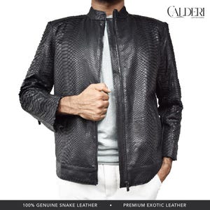 Op de afbeelding: Een zwarte slangenleren jas met ritssluiting. De jas heeft een textuurpatroon en een opstaande kraag. De tekst "CALDERI LEATHER GOODS" staat in de rechterbovenhoek. De onderkant van de afbeelding luidt "100% GENUINE SNAKE LEATHER" en "PREMIUM EXOTIC LEATHER."