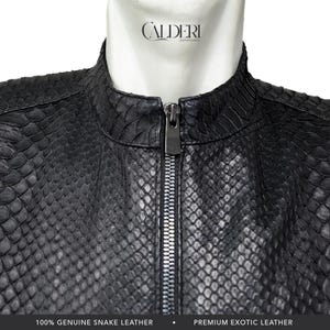 Könnte beinhalten: Schwarze Schlangenlederjacke mit Stehkragen und silbernem Reißverschluss. Die Jacke aus 100 % echtem Schlangenleder hat ein strukturiertes, schuppenartiges Muster. Der Markenname "CALDERI" ist oben sichtbar.
