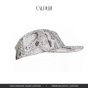 May include: A baseball cap crafted from genuine snake leather, showcasing a white and grey snakeskin design. The brand name "CALDERI" is visible at the top. The bottom of the image states "100% GENUINE SNAKE LEATHER" and "PREMIUM EXOTIC LEATHER."