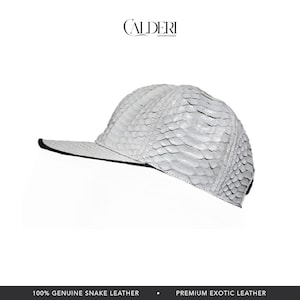 May include: A white, textured baseball cap made from genuine snake leather. The cap has a curved peak with a black edge and the brand name "CALDERI" at the top. A premium exotic leather product.