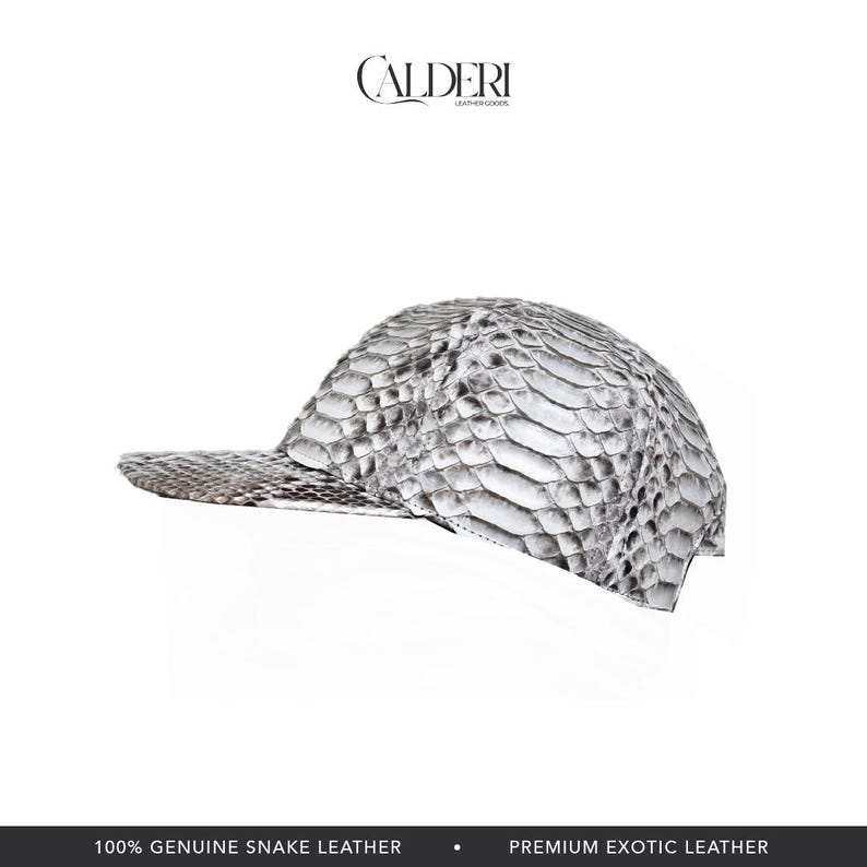 May include: A white and grey snake leather baseball cap. The cap has a curved peak and a textured pattern, with the brand name "CALDERI" at the top. The bottom of the image reads "100% GENUINE SNAKE LEATHER" and "PREMIUM EXOTIC LEATHER."