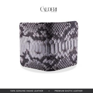 Op de afbeelding: Een bi-fold portemonnee gemaakt van echt slangenleer. De portemonnee heeft een grijs en zwart slangenpatroon. De merknaam "CALDERI" is bovenaan zichtbaar. De onderkant van de afbeelding luidt "100% ECHT SLANGENLEER" en "PREMIUM EXOTISCH LEER."