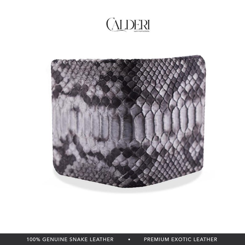 Op de afbeelding: Een bi-fold portemonnee gemaakt van echt slangenleer. De portemonnee heeft een grijs en zwart slangenpatroon. De merknaam "CALDERI" is bovenaan zichtbaar. De onderkant van de afbeelding luidt "100% ECHT SLANGENLEER" en "PREMIUM EXOTISCH LEER."