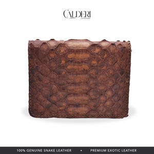Op de afbeelding: Een bruine, rechthoekige portemonnee van echt slangenleer. De portemonnee heeft een getextureerd, geschubd patroon en is gesloten. De merknaam "CALDERI" staat bovenaan. De onderkant van de afbeelding luidt "100% ECHT SLANGENLEER" en "PREMIUM EXOTISCH LEER."