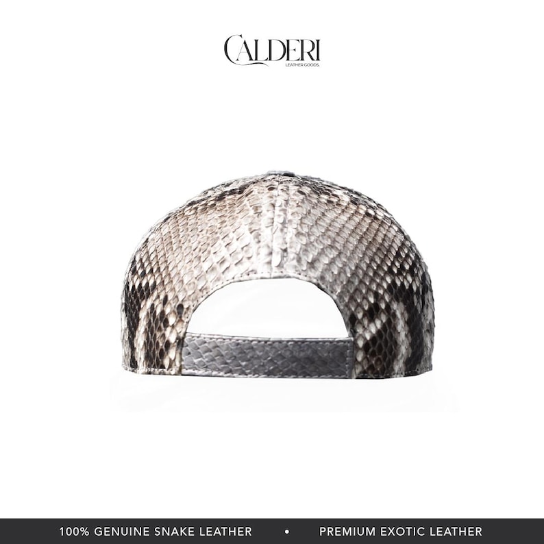 May include: A baseball cap crafted from genuine snake leather. The cap displays a snakeskin pattern in grey, black, and white. The brim is a solid grey colour. The brand name "CALDERI" is visible at the top. The text "100% GENUINE SNAKE LEATHER" is at the bottom.