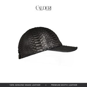 May include: A black baseball cap crafted from genuine snake leather. The cap has a textured, scale-like design and a curved peak. The brand name "CALDERI" is visible at the top. The bottom of the image reads "100% GENUINE SNAKE LEATHER" and "PREMIUM EXOTIC LEATHER."