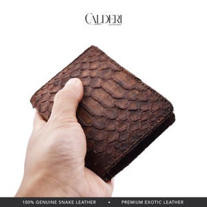 May include: A brown snake leather wallet with a textured, scale-like pattern. The wallet is held in a hand. The brand name "CALDERI" is displayed at the top. The wallet is made of genuine snake leather.