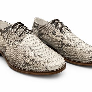 May include: A pair of lace-up oxford shoes featuring a textured snakeskin pattern in shades of grey, black, and off-white. The shoes have a low brown wooden heel, a rounded toe, and dark brown laces, showcasing a detailed reptilian scale design.