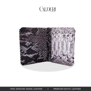 Op de afbeelding: Open dubbelgevouwen portemonnee van echt slangenleer. De portemonnee is open en onthult kaartsleuven en een gedeelte voor contant geld. De buitenkant heeft een zwart-grijs slangenpatroon. De merknaam "CALDERI" staat bovenaan.