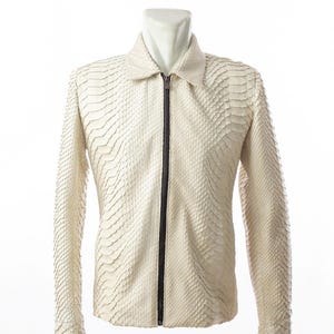 May include: A cream-coloured jacket featuring a textured, scale-like pattern across the entire surface. The garment includes a pointed collar, long sleeves with cuffs, and a dark brown vertical zip closure running down the centre front.
