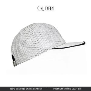 May include: White baseball cap with a snakeskin pattern. The cap has a curved brim with a black trim and a black adjustable strap. The brand name "CALDERI LEATHER GOODS" is at the top. The text at the bottom reads "100% GENUINE SNAKE LEATHER".