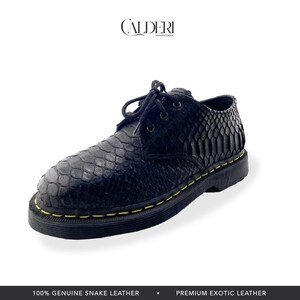 Op de afbeelding: Zwarte leren schoen met een slangenleerpatroon. De schoen heeft een vetersluiting, een ronde neus en een dikke zwarte zool met gele stiksels. De merknaam "CALDERI" is bovenaan zichtbaar. De tekst "100% GENUINE SNAKE LEATHER" staat onderaan.
