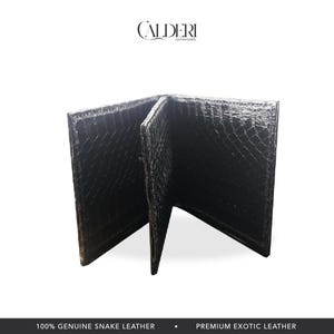 May include: Black bi-fold wallet made of genuine snake leather. The wallet is open, revealing the interior. The brand name "CALDERI" is displayed at the top. The bottom of the image reads "100% GENUINE SNAKE LEATHER" and "PREMIUM EXOTIC LEATHER."