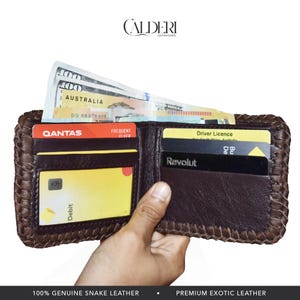 May include: Open brown leather wallet with woven edges, holding cash and cards. The wallet contains a Qantas frequent flyer card, a debit card, and a driver's license. The wallet is made of genuine snake leather.