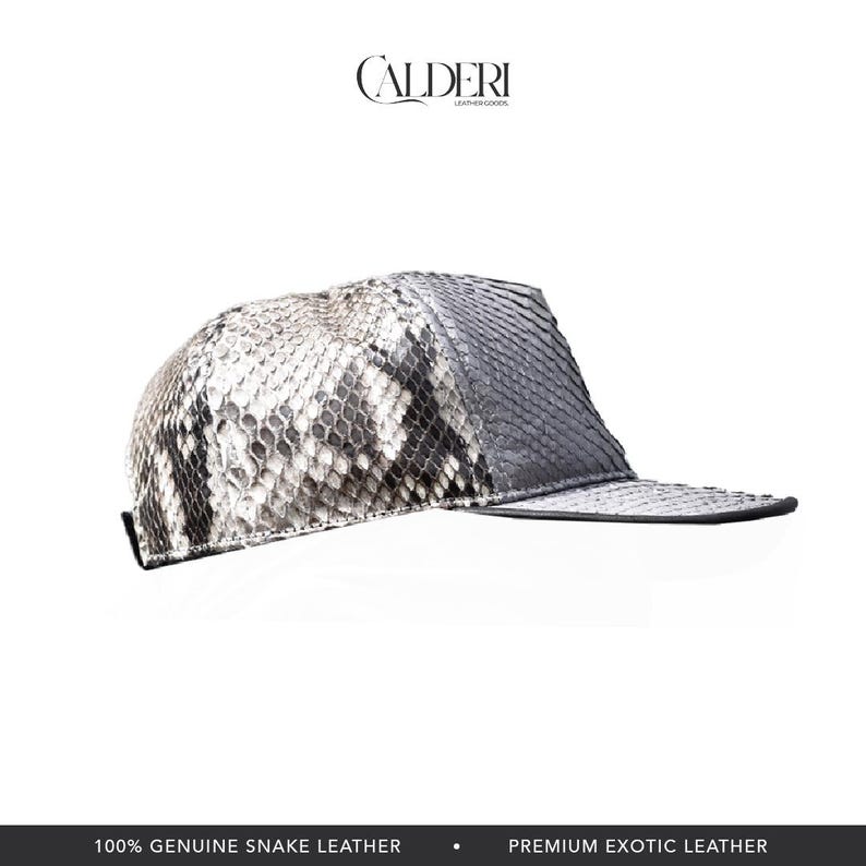 May include: A baseball cap crafted from genuine snake leather. The cap showcases a grey and white snakeskin pattern on the crown and a solid grey peak. The brand name "CALDERI" is displayed at the top. The text "100% GENUINE SNAKE LEATHER" is at the bottom.